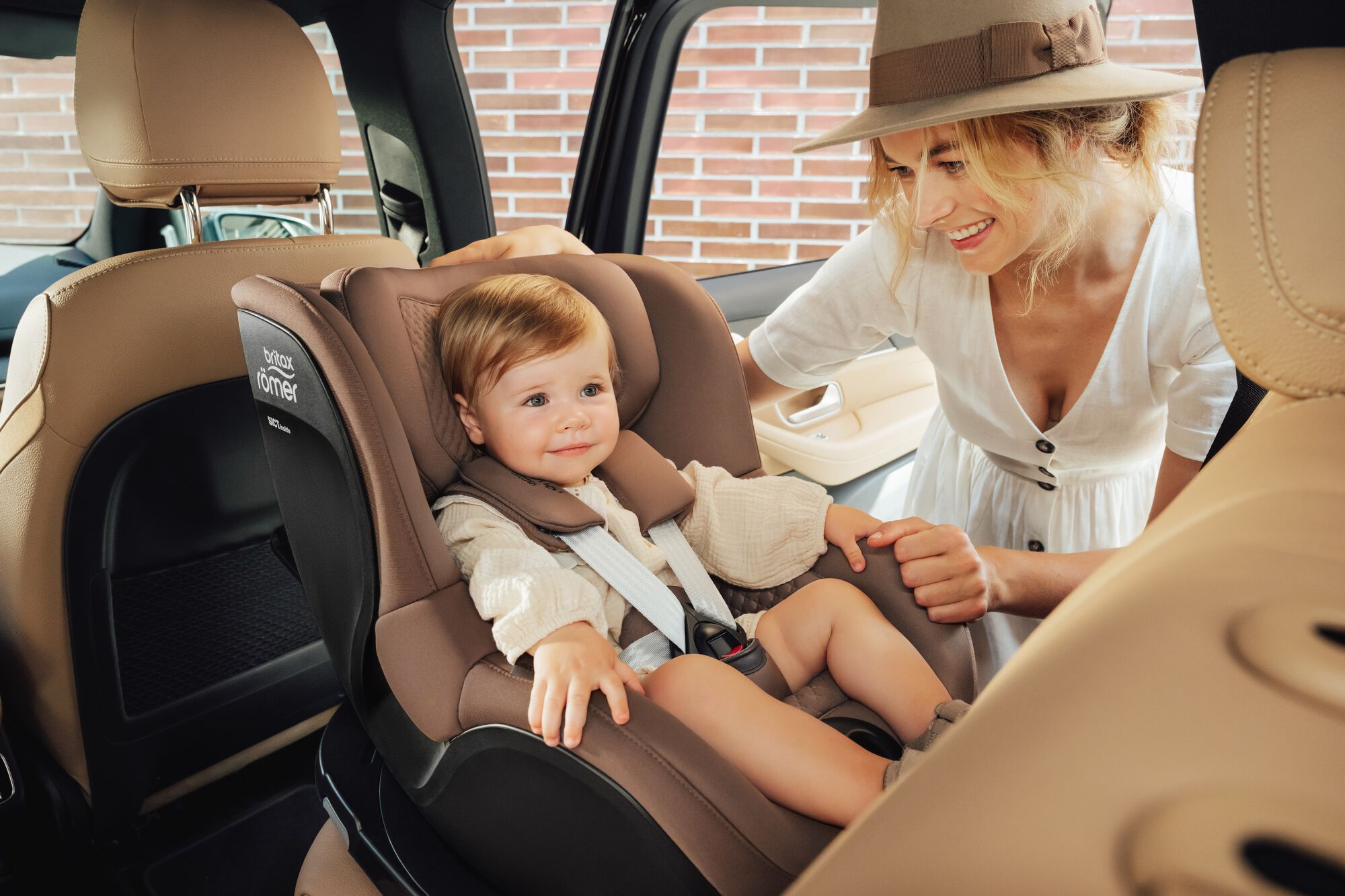 Maluch przypięty w foteliku samochodowym DUALFIX 5Z toddler car seat, z matką stojącą obok.